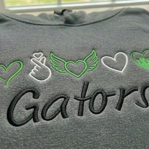 May include: A folded gray hoodie with embroidered designs. The design includes a green heart, a white hand gesture, a green heart with wings, a white heart, and a green heart with a dripping effect. The word "Gators" is embroidered in black.