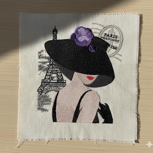 Pode incluir: Obra de arte bordada com uma mulher de chapéu preto com uma flor roxa, lábios vermelhos e luvas pretas. A Torre Eiffel e o texto "Paris Francaise 1982" também são bordados no tecido branco sujo.