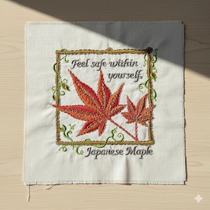 May include: Embroidered square textile featuring two orange and red Japanese maple leaves within a gold frame. The text "Feel safe within yourself" and "Japanese Maple" are embroidered in green.