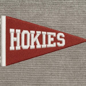 Puede incluir: Un banderín rojo con ribete blanco y la palabra "HOKIES" bordada en blanco. El banderín es triangular y tiene borlas blancas en el extremo. El fondo es una tela gris texturizada.