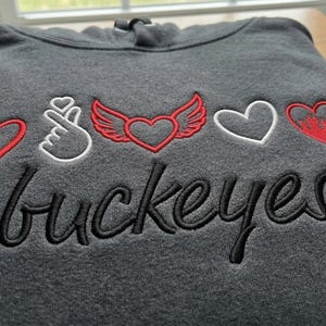 Könnte beinhalten: Dunkelgrauer Hoodie mit gestickten Herzen, einer Handgeste und dem Wort "buckeyes" in schwarzer Schrift. Das Design enthält rote und weiße Akzente und schafft ein visuell ansprechendes und thematisches Design.