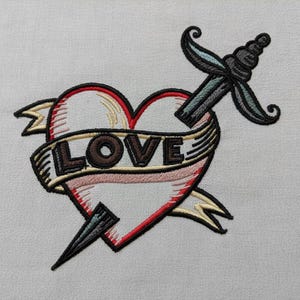 May include: Embroidered design featuring a heart pierced by a dagger, with a banner reading "LOVE". The heart is red with a white interior, outlined in black. The dagger and banner have black, green, and tan accents. The design is on a light gray fabric.