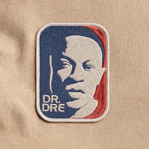 Puede incluir: Parche bordado con un retrato del Dr. Dre. El parche tiene un fondo azul con un borde rojo y las palabras "DR. DRE" en blanco. El parche es rectangular con esquinas redondeadas y está sobre un fondo de tela beige.