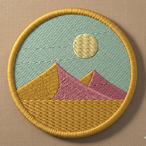 May include: Embroidered patch featuring a desert landscape with pyramids and a full moon. The design uses yellow, pink, and blue-green thread, with a yellow border. The patch is circular and appears to be made of fabric.