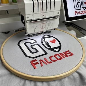 May include: An embroidery machine in operation, stitching a design onto grey fabric held in a wooden hoop. The design features the words "GO FALCONS" in black and red thread, and a football graphic with a red heart.