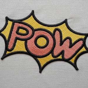 May include: Embroidered patch with the word "POW" in a comic book style. The word is in a coral color with a black outline, set against a yellow burst shape with a black border. The patch is on a light gray fabric.
