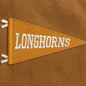 Könnte beinhalten: Gestickter Wimpel in Dreiecksform mit dem Wort "LONGHORNS" in weißen Großbuchstaben. Der Wimpel ist goldgelb mit weißen Fransen. Der Hintergrund ist ein strukturierter, brauner Stoff.