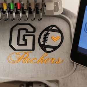 May include: Close-up of an embroidery machine stitching a gray fabric with the words "GO Packers". The design includes a black and yellow football and a yellow heart.