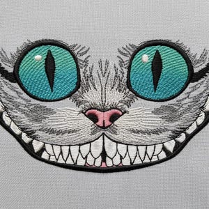 May include: Embroidered patch of the Cheshire Cat's face. The cat has a gray and white face, large teal eyes, and a pink nose. Its mouth is open, revealing sharp white teeth. The patch is set against a light grey background.