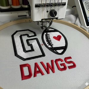 Op de afbeelding: Een borduurmachine borduurt een ontwerp op witte stof. Het ontwerp bevat de woorden "GO DAWGS" in rode draad, met een zwart-witte voetbal en een rood hart. De letter "G" is zwart omlijnd.