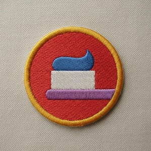 May include: Embroidered patch featuring a toothbrush with toothpaste. The design includes a red circle with a yellow border, a white toothbrush, purple handle, and blue toothpaste. The patch is likely for clothing or accessories.