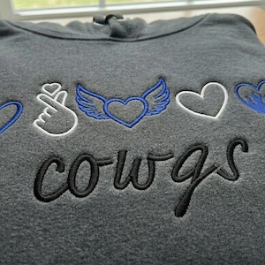 Puede incluir: Una sudadera con capucha gris carbón doblada con diseños bordados. Los diseños incluyen un corazón azul, un gesto con la mano, un corazón con alas, un corazón blanco y un corazón azul con efecto goteo. La palabra "cowgs" está bordada en escritura negra.