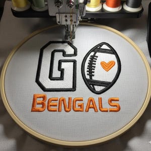 Puede incluir: Un bastidor de bordado con las palabras "GO BENGALS" bordadas en hilo negro y naranja. La letra "G" está delineada en blanco y negro, y junto a ella un balón de fútbol americano con un corazón. El bordado está sobre tela gris claro.