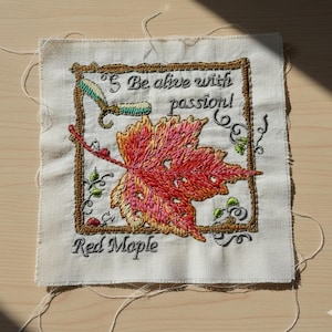 May include: Embroidered square of fabric featuring a red maple leaf with gold and pink accents. The text "Be alive with passion!" and "Red Maple" are embroidered in black. The design is framed with a brown border.