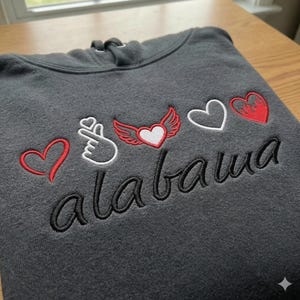 Pode incluir: Moletom cinza escuro com corações bordados, um gesto de mão e a palavra "alabama". Os corações são vermelhos e brancos, e um tem asas. O moletom tem capuz com cordão.