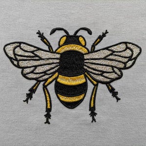 May include: Embroidered bee design in black, yellow, and silver on a light gray background. The bee has detailed wings and body stripes, with delicate antennae and legs. The embroidery is finely stitched, creating a textured effect.