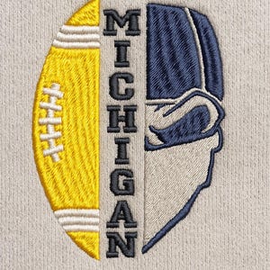 Puede incluir: Diseño bordado con un balón de fútbol amarillo y un casco azul, dividido verticalmente. La palabra "MICHIGAN" está bordada en letras mayúsculas negras en el centro. El diseño está sobre un fondo gris claro.