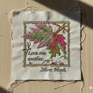 May include: Embroidered square of fabric featuring a silver maple leaf in shades of pink, green, and orange. The text "Love one another" and "Silver Maple" are embroidered below the leaf, framed by a gold border.