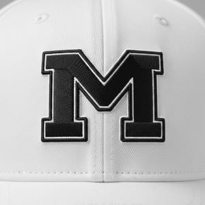 May include: A white baseball cap featuring a large, black letter "M" with a white outline. The letter is embroidered on the front of the cap. The cap has a curved brim and a structured crown.