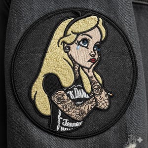 May include: Embroidered patch featuring a distressed Alice in Wonderland design. Alice has blonde hair, blue eyes, and tattoos. She is wearing a black shirt with the text "K Dane" and "Tennessee". The patch is circular and set against a dark gray background.