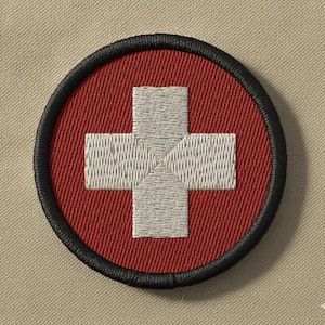 May include: Embroidered patch with a white cross on a red circular base, edged with a black border. The design suggests a medical or first aid theme, with a textured fabric finish.