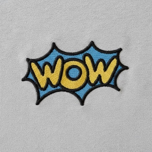 Pode incluir: Patch bordado com a palavra "WOW" em amarelo, contornado em preto, sobre um fundo azul claro. O patch tem a forma de um balão de banda desenhada e está sobre um tecido cinzento claro.