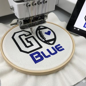 Può includere: Un telaio da ricamo con le parole "GO BLUE" ricamate con filo nero e blu. Il disegno include un pallone da football con un cuore al centro. La macchina sta ricamando.