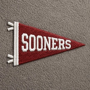 May include: A maroon pennant with the word "SOONERS" embroidered in white. The pennant has a white border and decorative white fringe. The background is a textured gray fabric.