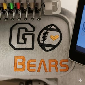 May include: Embroidered design on light gray fabric. The design features the words "GO BEARS" in black and orange thread. A football graphic with a yellow heart is also included. The image is taken during the embroidery process.