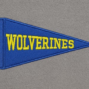 Pode incluir: Uma flâmula azul com texto bordado amarelo que diz "WOLVERINES". A flâmula tem franjas amarelas e está sobre um fundo cinza. Este item é provavelmente uma lembrança de equipe ou escolar.
