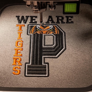 Puede incluir: Una máquina de bordar cosiendo un parche con las palabras "WE ARE TIGERS". El parche presenta una gran letra "P" negra con un diseño de cabeza de tigre naranja en la parte superior. El texto "TIGERS" está alineado verticalmente a la izquierda en naranja.