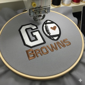 May include: Embroidered design on gray fabric within a wooden hoop. The design features the words "GO BROWNS" in white and brown thread, along with a football graphic with a heart. The embroidery is being done by a machine.