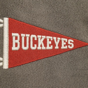 Puede incluir: Un banderín rojo con la palabra "BUCKEYES" bordada en blanco. El banderín tiene un borde blanco y detalles blancos en forma de flecos en la parte superior. El fondo es una tela gris texturizada.