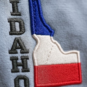 Op de afbeelding: Geborduurde patch met de staat Idaho in rood, wit en blauw. Het woord "IDAHO" is verticaal in zwart gestikt. De patch bevindt zich op een lichtblauwe stoffen achtergrond.