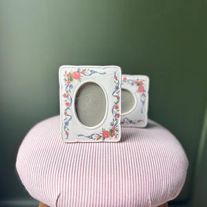 Puede incluir: Dos marcos de fotos ovalados blancos con diseños florales y de cintas. Los marcos tienen un borde festoneado y están decorados con rosas rosas, cintas azules y hojas verdes. Se encuentran sobre un taburete a rayas rosas y blancas.