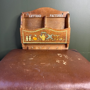 May include: A wooden wall-mounted mail organiser with two compartments labelled "LETTRES" and "FACTURES" in white text. The front panel features a painted illustration of a teapot, fruit, and utensils, with four small metal hooks attached below.