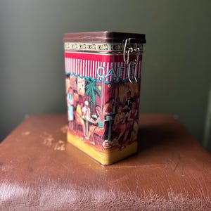 Puede incluir: Una lata de metal rectangular con tapa marrón y cierre plateado. La lata presenta una colorida ilustración de una escena de café con gente, palmeras y la palabra "cafe" repetida en la parte superior. La lata está sobre una superficie marrón.