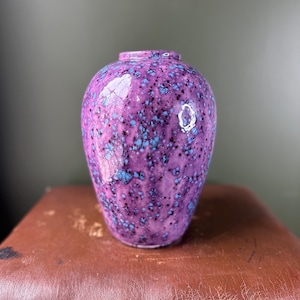 May include: A ceramic vase with a rounded body and a narrow opening, featuring a glossy purple glaze speckled with vibrant blue and dark black spots throughout the surface, creating a textured appearance with a reflective finish on the exterior.