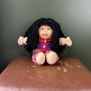 May include: A doll with black yarn hair, a light skin tone, and a soft fabric body. The doll wears a purple shirt with a red collar and a red button-down placket, paired with colorful floral print pants featuring pink, green, and blue patterns.