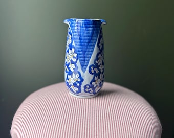 May include: A blue and white ceramic vase with a floral design. The vase has a unique shape with a flared top and is decorated with white flowers and blue accents. The vase is approximately 6 inches tall.