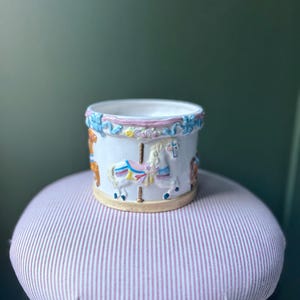Op de afbeelding: Witte keramische carrouselplanter met kleurrijke paarden en decoratieve rand. De cilindrische planter staat op een roze en wit gestreepte kruk. De carrouselpaarden zijn in tinten blauw, roze en geel.
