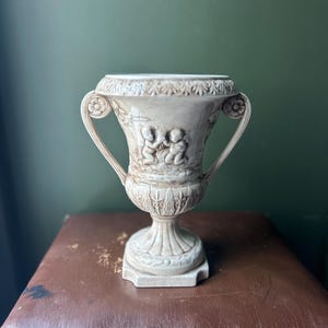 Op de afbeelding: Een crèmekleurige, decoratieve urn met twee handvatten. De urn heeft een gedetailleerd reliëf van cherubijnachtige figuren en bloemmotieven. De basis is vierkant en de urn staat tegen een donkergroene achtergrond.