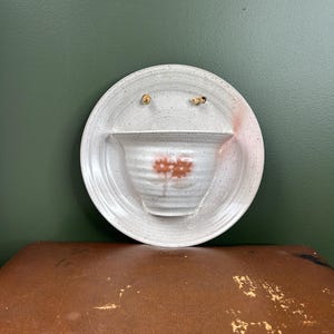 May include: A circular ceramic wall planter with a speckled off-white glaze and a ribbed bowl section. The piece features a painted orange flower motif on the front and two small holes at the top for hanging with a light-coloured cord.