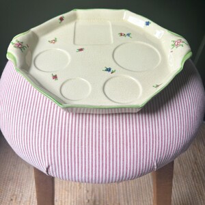 Könnte beinhalten: Ein achteckiges Vintage-Serviertablett in Creme, verziert mit kleinen Blumenmustern in Rot, Rosa und Blau. Das Tablett hat eine grüne Umrandung und vier kreisförmige Vertiefungen für Tassen. Es liegt auf einem gestreiften Hocker mit rot-weißen Streifen.