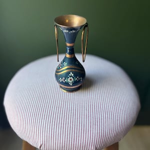 May include: A small, dark blue vase with a gold-colored rim and handles. The vase is decorated with gold and white floral patterns. It sits on a striped, round stool with wooden legs.