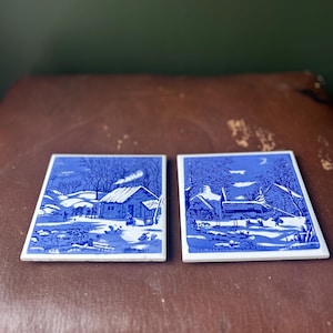 May include: Two square blue and white ceramic coasters depicting winter scenes. Each coaster shows a detailed illustration of a cabin, trees, and figures in a snowy landscape. The coasters have a white border.