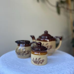 Op de afbeelding: Een vintage keramische theeset met een theepot, een melkkan en een pot met deksel. De set heeft een crèmekleurige basis met bruine bloemmotieven en donkerbruine accenten. De theepot heeft een handvat en een tuit, en de pot heeft een kleine knop op het deksel.