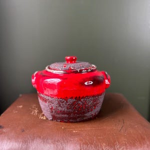 Könnte beinhalten: Ein kleiner, roter Keramikbehälter mit Deckel. Der Behälter hat einen strukturierten, dunkelbraunen Boden und eine glänzend rote Oberseite. Der Deckel ist ebenfalls dunkelbraun mit einem kleinen roten Knopf. Zwei kleine Griffe befinden sich an den Seiten.