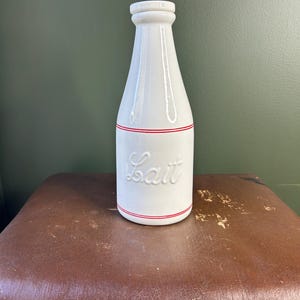 May include: A white ceramic milk bottle with a narrow neck and a matching lid. The bottle features the word Lait embossed in cursive script on the front, accented by two thin horizontal red stripes encircling the body above and below the text.