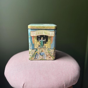 Può includere: Scatola rettangolare con un'illustrazione colorata di una scena di fattoria, tra cui un contadino, polli e la scritta "Jersey Milk". La scatola è posizionata su uno sgabello rotondo con un motivo a righe rosse e bianche.
